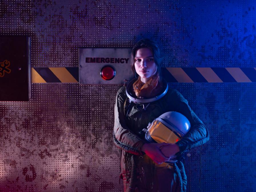 En kvinde iført rumdragt står foran en rød emergency knap